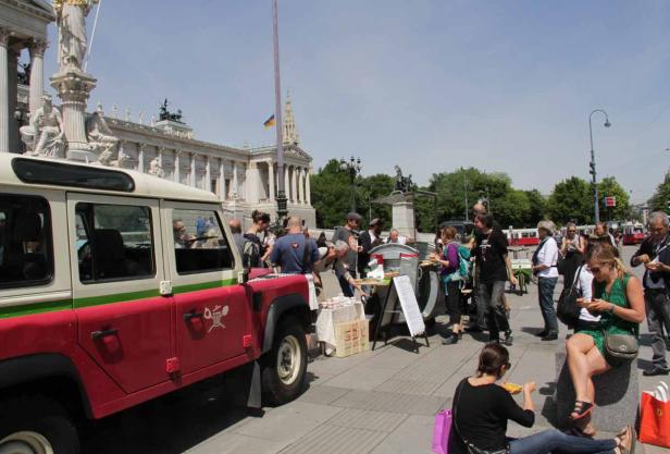 Ein Imbissstand vor dem Wiener Parlament mit Menschen und einem roten Geländewagen.