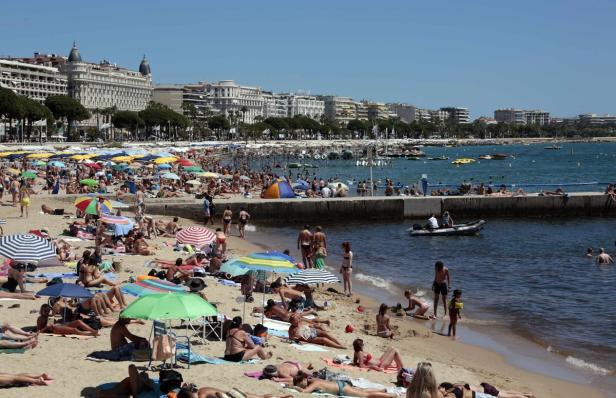 Ein belebter Strand mit vielen Menschen und bunten Sonnenschirmen vor einer Stadtkulisse.