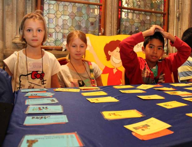 Drei Kinder sitzen an einem Tisch mit Karten und einem Cartoon im Hintergrund.