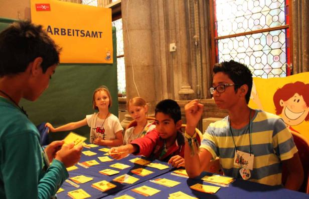 Kinder spielen ein Spiel mit Kärtchen vor einem Schild mit der Aufschrift „Arbeitsamt“.