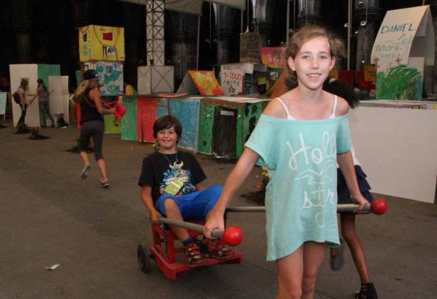 Ein Mädchen schiebt einen Jungen in einem roten Wagen vor einer bunten Kulisse.