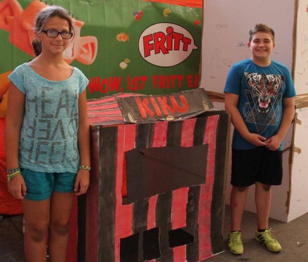 Ein Mädchen mit Brille und ein Junge stehen neben einem bemalten Kiosk mit der Aufschrift „KIKU“.