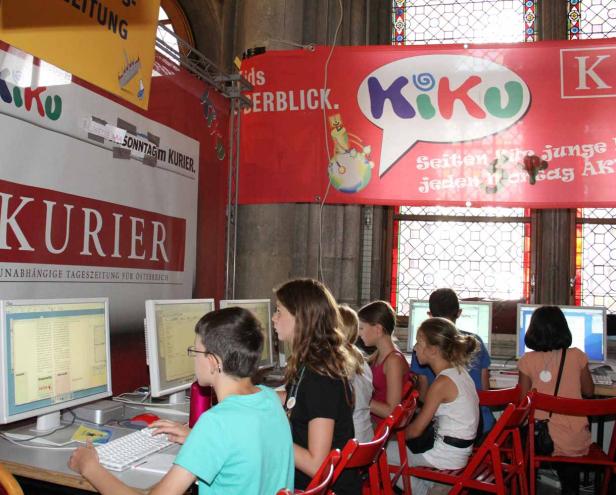 Kinder sitzen an Computern unter Bannern der Zeitung „Kurier“ und der Kinderseite „Kiku“.