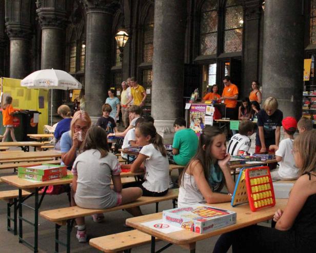 In einer Halle spielen Kinder und Erwachsene Brettspiele an Biertischen.