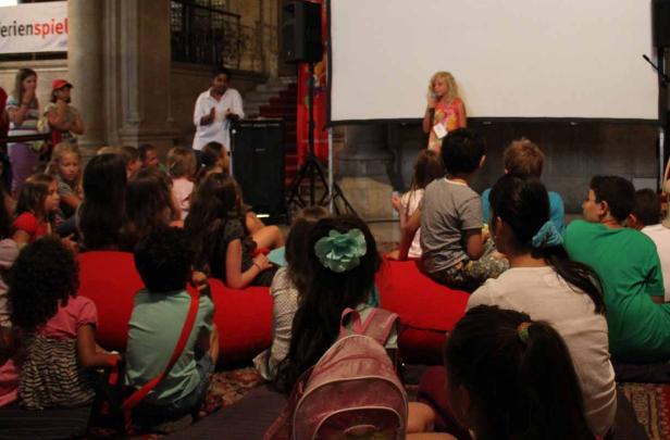 Eine Gruppe Kinder sitzt auf roten Sitzsäcken und schaut einer jungen Rednerin zu.