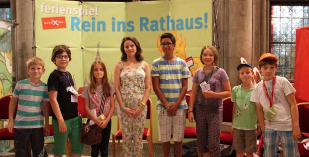 Eine Gruppe von Kindern steht vor einem Banner mit der Aufschrift „Ferienspiel Rein ins Rathaus!“.