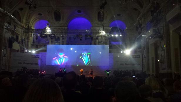 Eine Bühnenaufführung mit Tänzern in leuchtenden Kostümen vor einem großen Publikum.