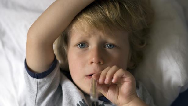 Ein blonder Junge liegt mit einem Thermometer im Mund im Bett.