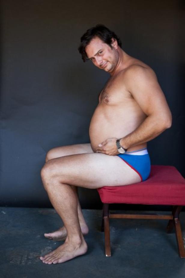 Ein Mann sitzt mit freiem Oberkörper und blauer Unterhose auf einem roten Hocker.