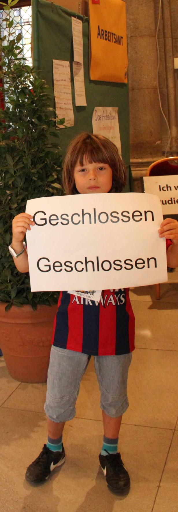 Ein Kind hält ein Schild mit der Aufschrift „Geschlossen“ vor einem Aushang des Arbeitsamtes.