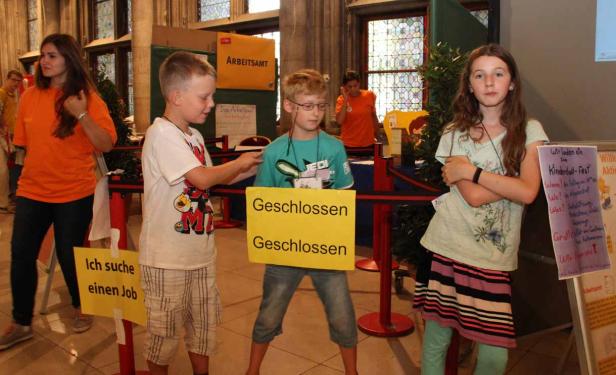 Kinder stehen vor einem Stand mit Schildern, auf denen „Geschlossen“ und „Ich suche einen Job“ steht.