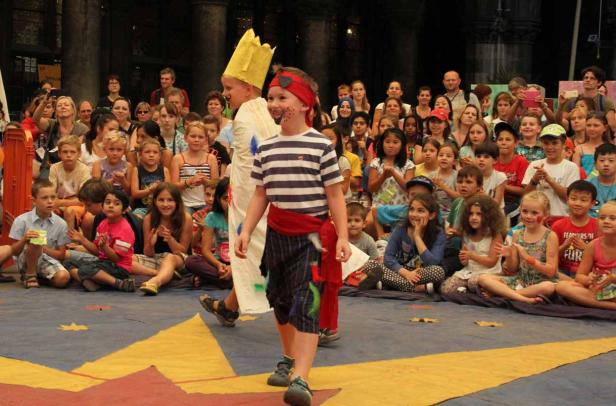 Kinder in Kostümen führen ein Theaterstück vor einem Publikum auf.