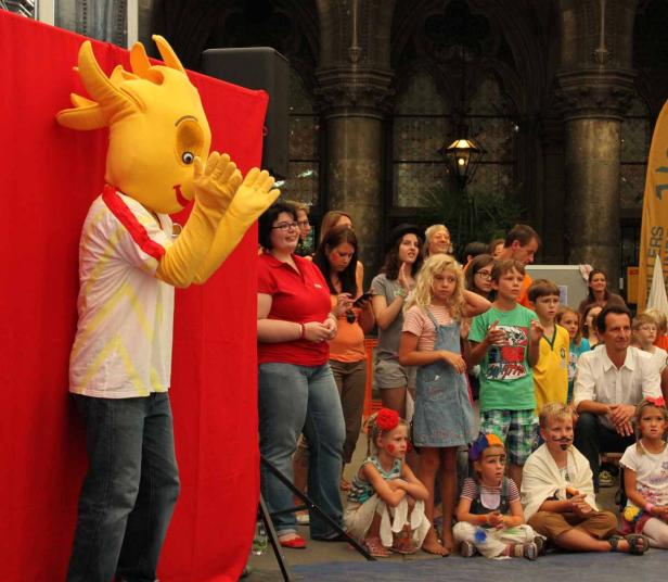 Ein gelbes Maskottchen klatscht vor einer Gruppe von Kindern und Erwachsenen.