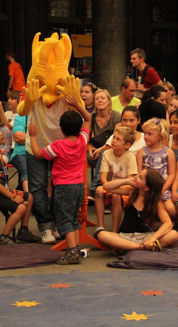Ein Kind interagiert mit einem gelben Maskottchen vor einer Gruppe von Zuschauern.