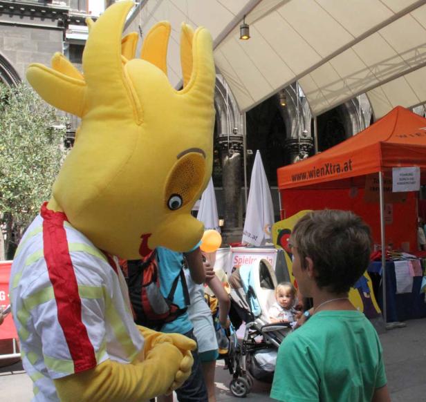 Ein gelbes Maskottchen von WienXtra interagiert mit einem Kind auf einer Veranstaltung.