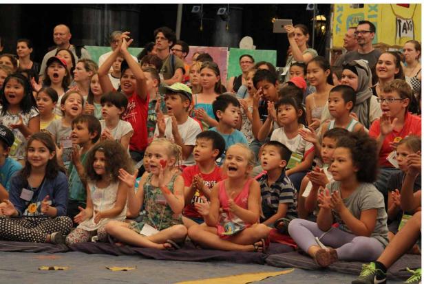 Eine Gruppe von Kindern und Erwachsenen sitzt auf dem Boden und klatscht begeistert.