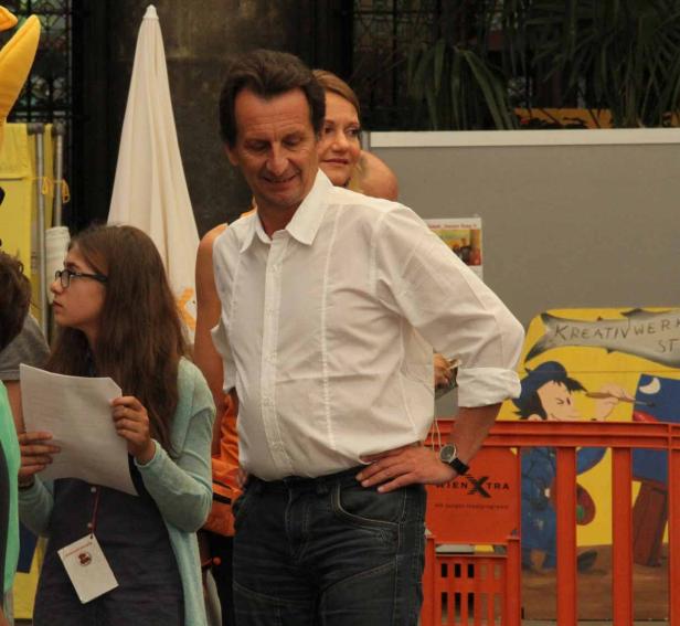 Ein Mann in Hemd und Jeans steht mit verschränkten Armen vor einer Gruppe von Menschen.