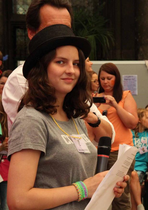 Eine junge Frau mit Hut hält ein Mikrofon und ein Blatt Papier in der Hand.