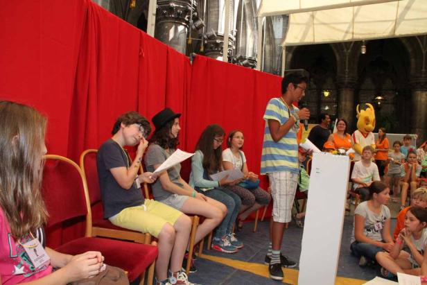 Ein junger Mann spricht vor einer Gruppe von Kindern in einem Raum mit rotem Vorhang.