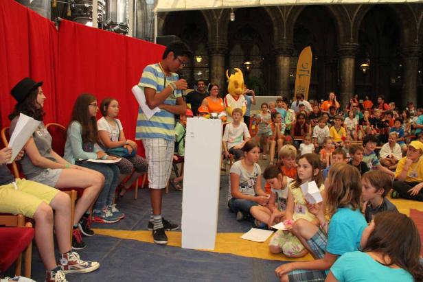 Ein junger Mann hält eine Rede vor einer Gruppe von Kindern und einem Maskottchen.