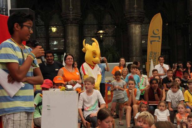 Eine Gruppe von Kindern und Erwachsenen versammelt sich um einen Redner und ein Maskottchen.