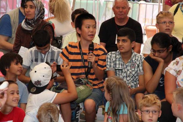 Ein Junge spricht mit einem Mikrofon vor einer Gruppe von Kindern und Erwachsenen.