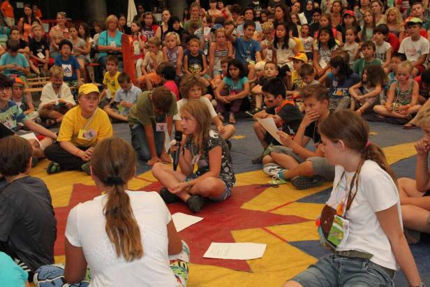 Eine Gruppe Kinder sitzt auf dem Boden und hört einer jungen Sprecherin zu.