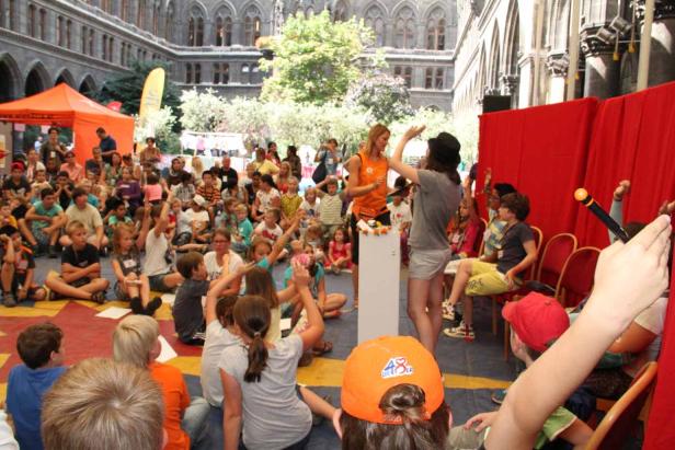 Eine Gruppe von Kindern sitzt und steht auf einem Platz und hört aufmerksam zu.