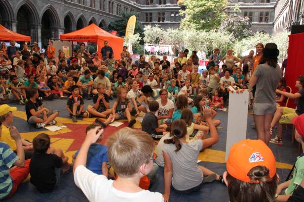 Eine Gruppe Kinder sitzt auf dem Boden und schaut einer Frau mit Zylinder zu.
