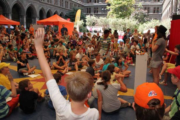 Eine Gruppe Kinder sitzt auf dem Boden und hört einer Frau zu, die auf einer Bühne steht.