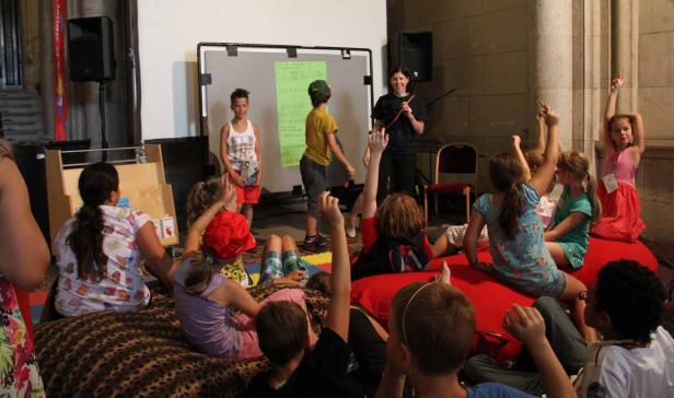 Eine Gruppe Kinder sitzt auf Kissen und hört einer Frau zu, die vor einer Tafel steht.