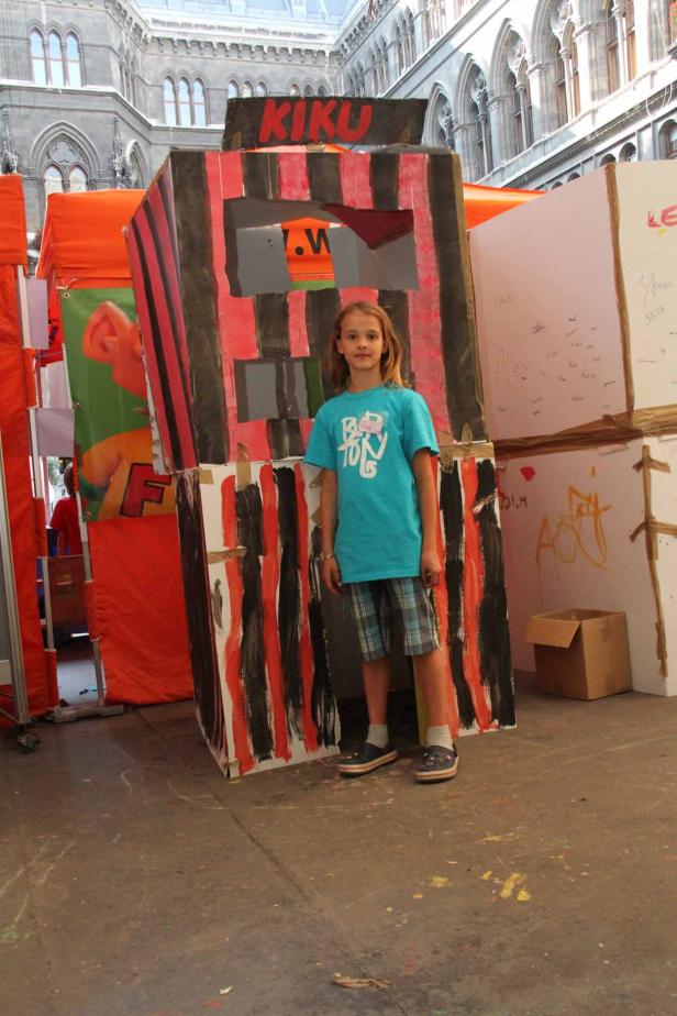 Ein junges Mädchen steht vor einem bemalten Papphaus mit dem Schriftzug „KIKU“.