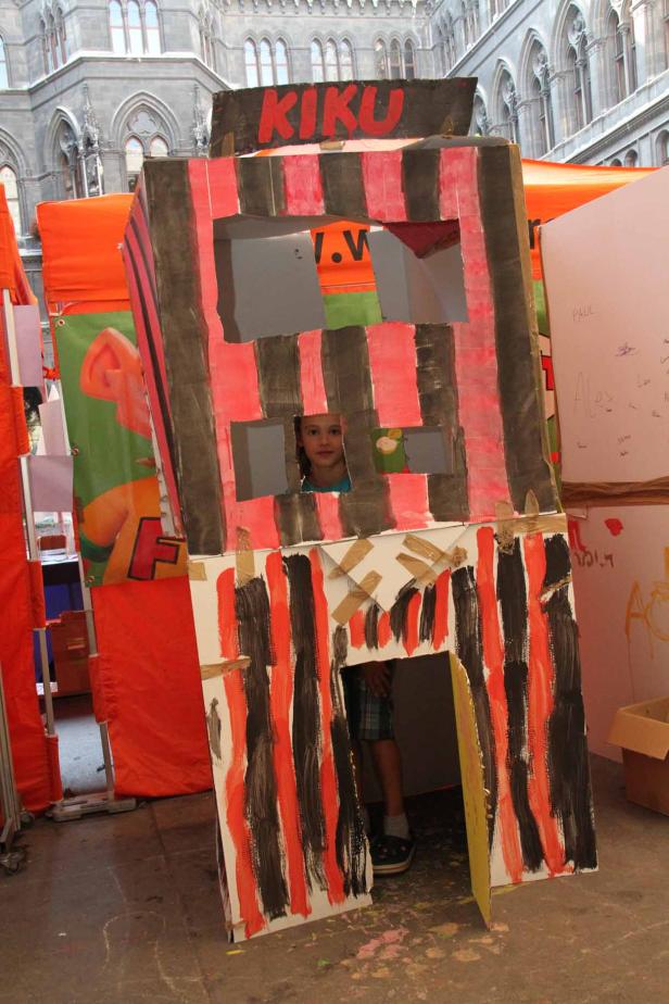 Ein Kind schaut aus einem selbstgebauten Kiosk mit der Aufschrift „KIKU“.