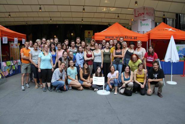 Eine große Gruppe von Menschen posiert vor orangefarbenen Zelten von WienXtra.