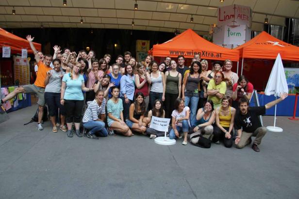 Eine große Gruppe von Menschen posiert vor orangefarbenen Zelten für ein Foto.