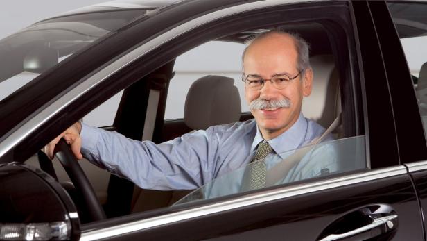 Ein lächelnder Mann mit Schnurrbart sitzt am Steuer eines schwarzen Autos.