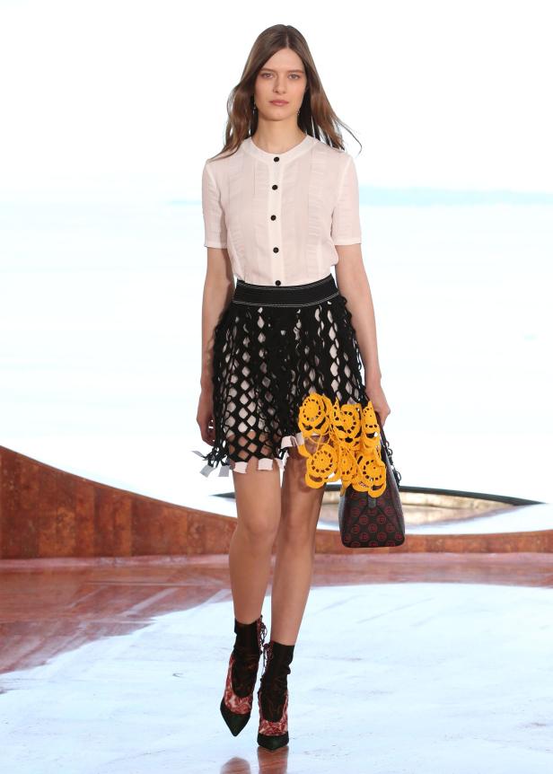 Eine Frau auf dem Laufsteg mit einem weißen Hemd, einem schwarzen Rock und einer Handtasche von Dior.