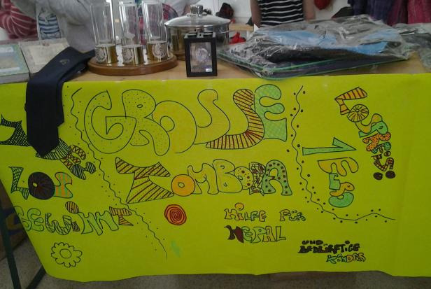 Ein Tisch mit Gegenständen und einem gelben Plakat für eine Tombola zur Hilfe für Nepal.