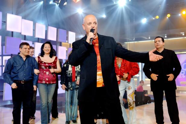 Ein Mann mit Mikrofon steht auf einer Bühne, umgeben von anderen Personen.