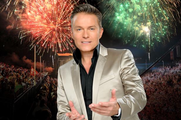 Ein Mann in einem Anzug steht vor einer Menschenmenge und Feuerwerk im Hintergrund.