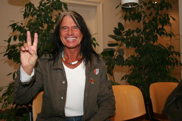 Ein lächelnder Mann mit langen Haaren zeigt das Victory-Zeichen.