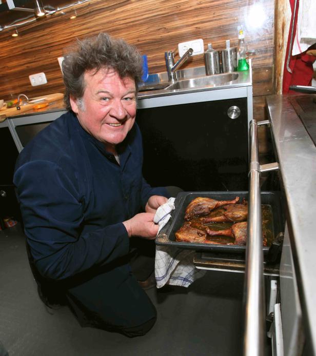 Ein lächelnder Koch kniet in einer Küche und präsentiert eine Pfanne mit Entenkeulen.