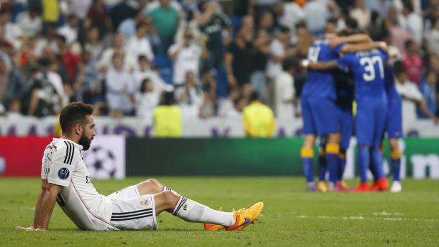 Ein enttäuschter Fußballspieler sitzt auf dem Rasen, während das gegnerische Team im Hintergrund jubelt.