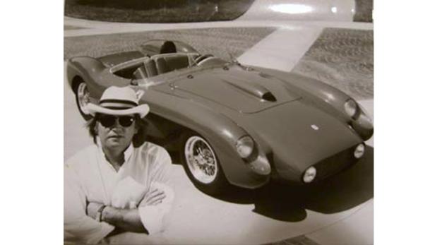 Ein Mann mit Hut posiert neben einem klassischen Ferrari-Rennwagen.