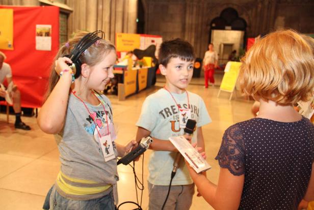 Zwei Kinder interviewen eine Person mit Mikrofonen auf einer Veranstaltung.