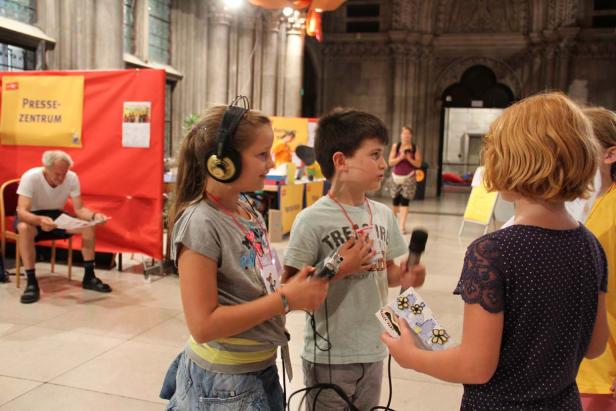 Zwei Kinder interviewen ein anderes Kind mit Mikrofonen in einem Raum.