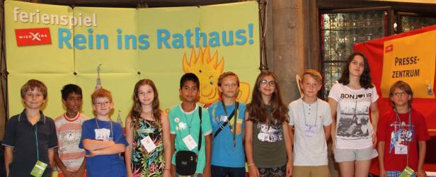 Eine Gruppe von Kindern steht vor einem Banner für das „Ferienspiel Rein ins Rathaus!“.