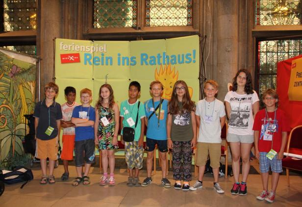 Eine Gruppe von Kindern steht vor einem Banner mit der Aufschrift „Ferienspiel Rein ins Rathaus!“.