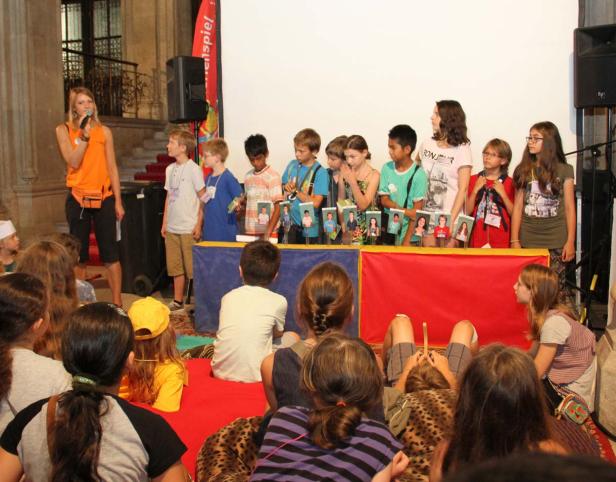Eine Frau moderiert eine Veranstaltung, bei der Kinder auf einer Bühne stehen.