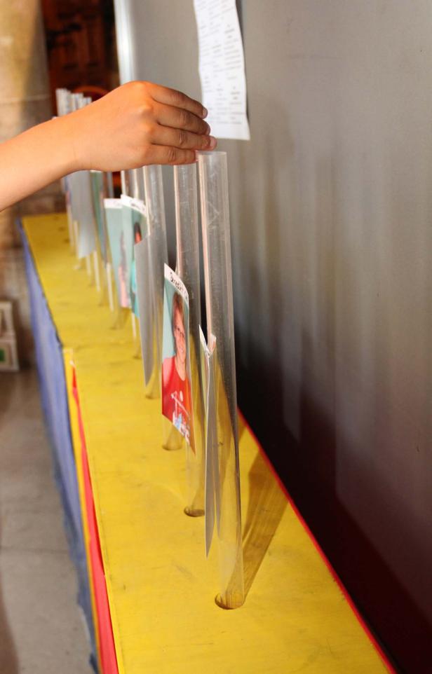 Eine Hand berührt eine Reihe von Fotos, die in transparenten Röhren auf einem gelben Tisch stehen.